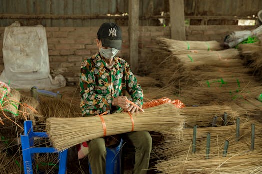 A masked artisan skillfully ties broomsticks in an indoor market setting.