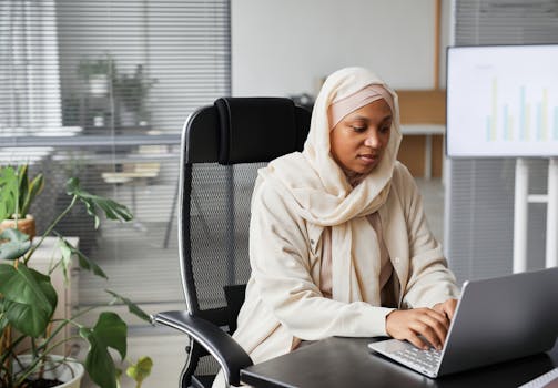 Professional Muslim woman in hijab working on laptop in modern office setting, demonstrating focus and dedication.