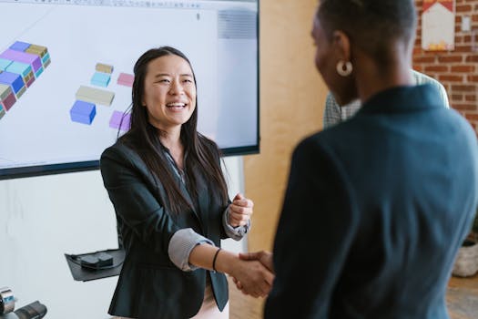 Two women shaking hands in a modern office environment with a presentation screen.