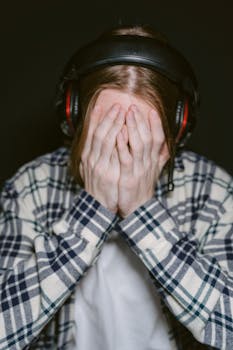 A young gamer covers face with hands in frustration, wearing headphones and plaid shirt in a dimly lit room.